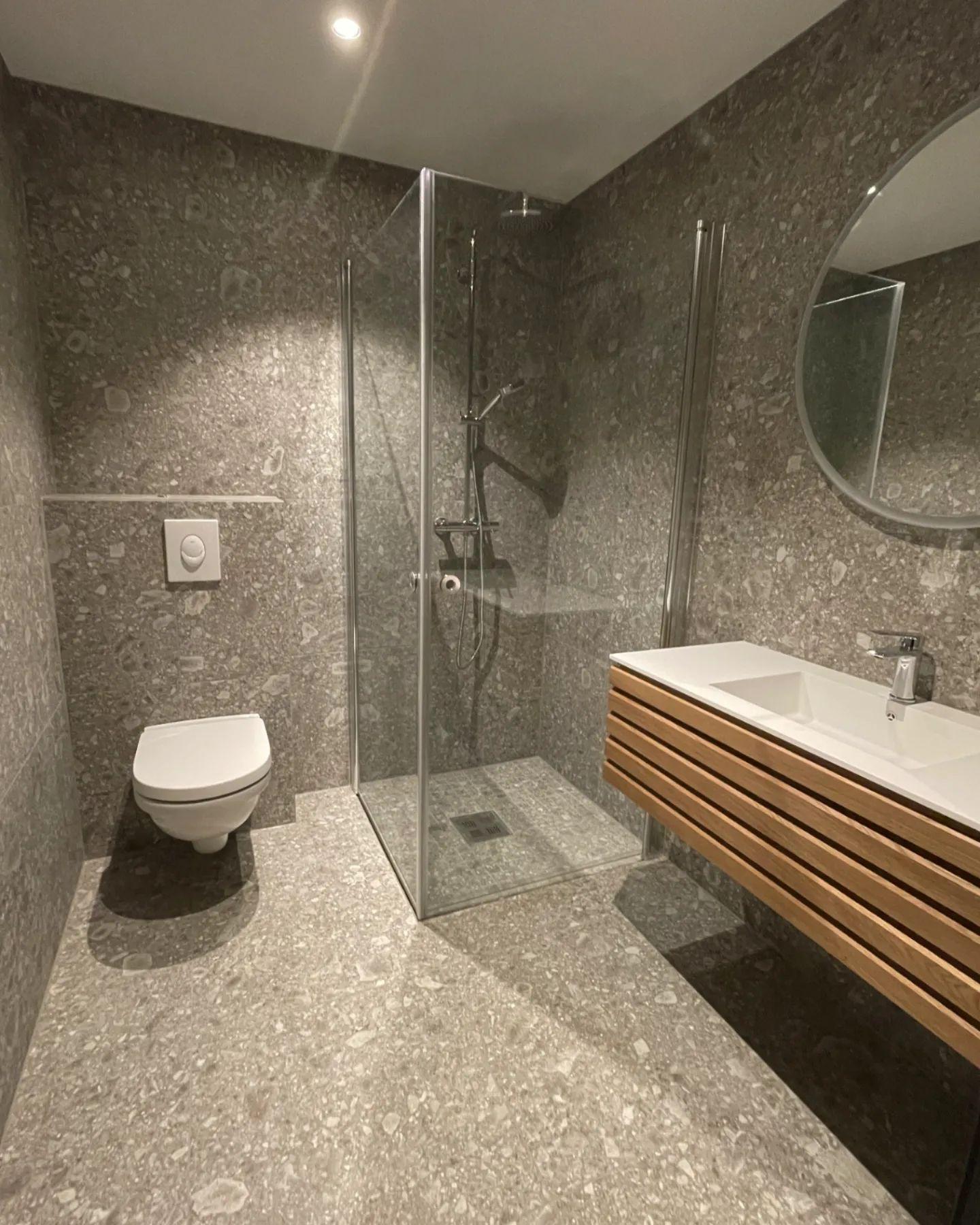 A bathroom with stone-look tiles, a walk-in shower behind glass and a wall-hung toilet